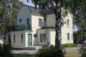 The yellow hostel house with a mansard roof, green door and balcony, behind a birch tree