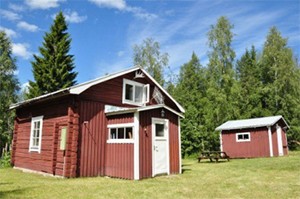 Die rote Angelhütte mit weißen Ecken und einem kleinen Schuppen daneben, umgeben von Fichtenwald